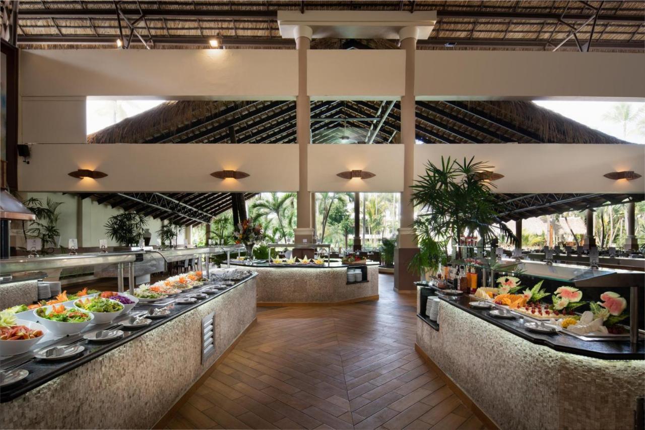 Impressive Punta Cana Hotel Exterior photo The photo depicts a spacious and elegantly designed restaurant interior featuring a buffet setup. There are several low tables lined with an array of colorful dishes that include fresh salads, fruits, and various entrees. The dining area is illuminat