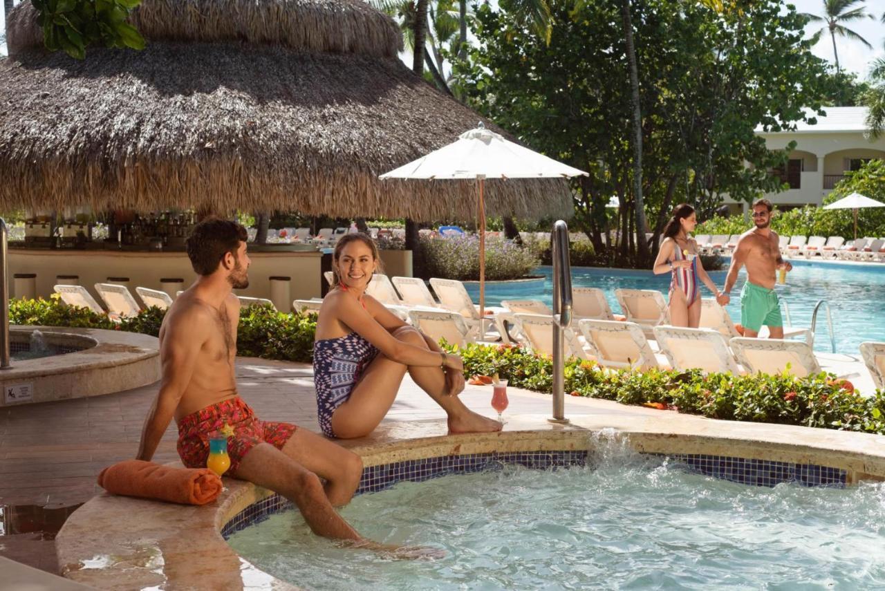 Impressive Punta Cana Hotel Exterior photo The photo shows a tropical pool scene. In the foreground, a man and woman are relaxing in a hot tub. The man is shirtless, wearing colorful swim trunks, and he has a drink in his hand. The woman is sitting beside him, wearing a stylish swimsuit. 

In