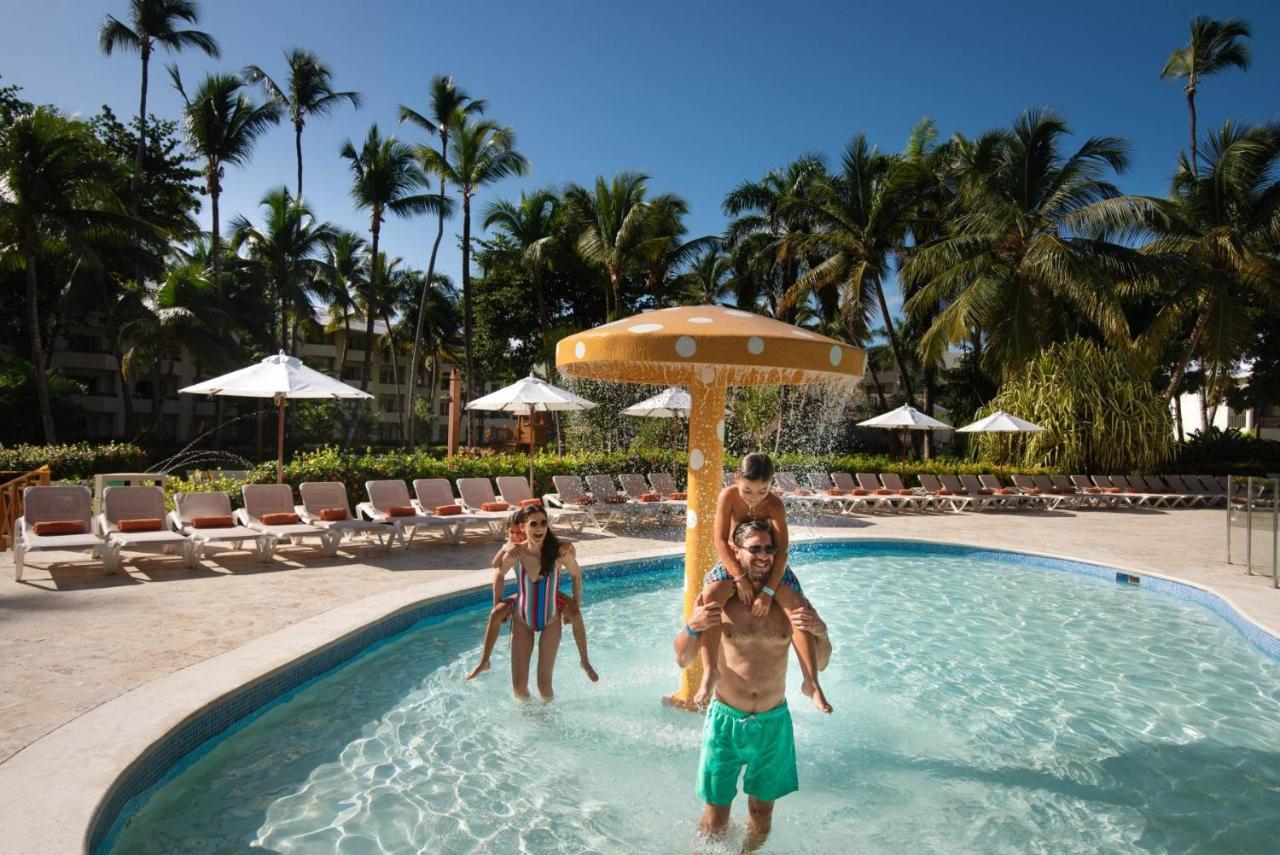 Impressive Punta Cana Hotel Exterior photo The photo shows a lively pool scene under a clear blue sky. In the foreground, there are two children playing; one is sitting on the shoulders of an adult. They are enjoying the water and the sunny atmosphere. Nearby, there is a water feature resembl