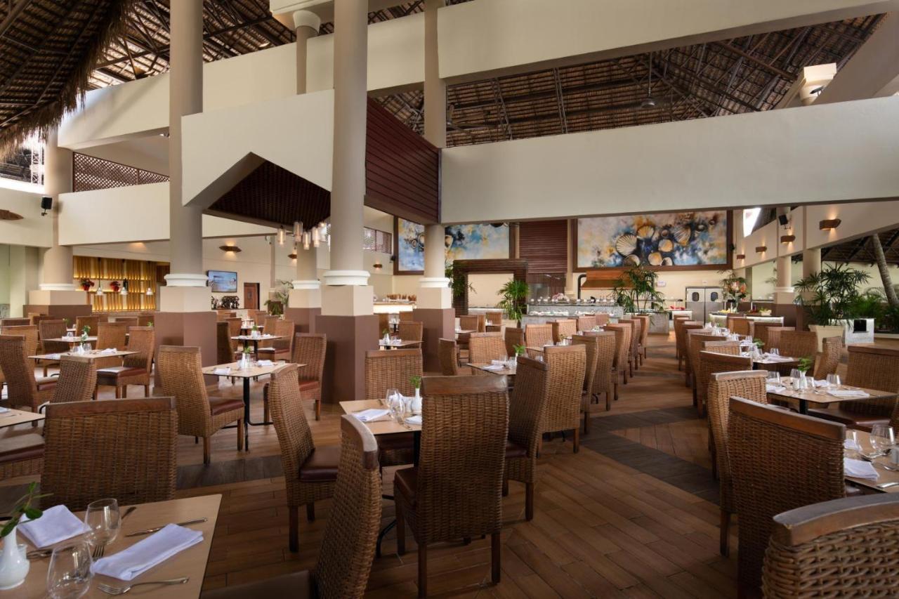 Impressive Punta Cana Hotel Exterior photo The photo shows an indoor restaurant with a spacious and modern design. The dining area features numerous tables set with cutlery and napkins. There are wicker chairs surrounding the tables, giving a comfortable and casual atmosphere. The ceiling is 